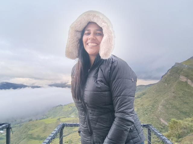 PauletteLoveX in a black puffy jacket with a fluffy pink hood, smiling at the camera, standing on a metal fence with misty green hills and mountains in the background.