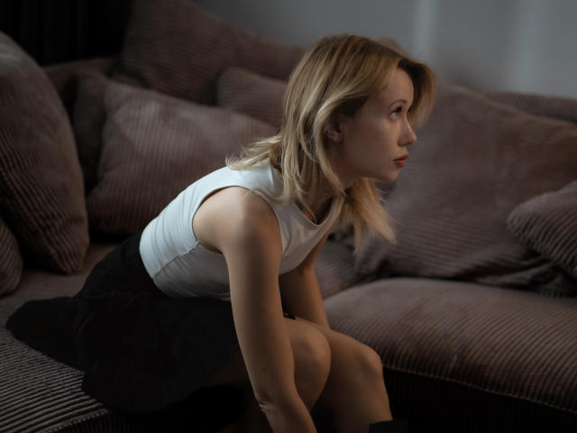 MisEmmaWonder with blonde hair, white sleeveless top, black skirt, and knee socks, kneeling on a gray, ribbed couch, looking to the side, with a serious expression.