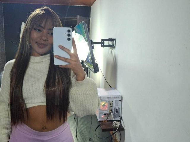 AmelieDuvall with long brown hair, white crop top, and pink pants, holding a phone in front of a mirror, showing her belly button and smiling.