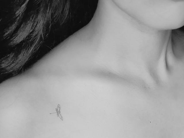 OranaSub's neck with a small fly tattoo on the upper chest. Her skin is smooth, and her dark hair is partially visible. Black and white photo.