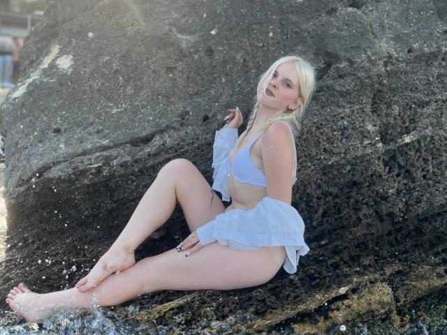 LaPulia, met bleke huid, blond haar en kleine borsten, draagt een witte beha en een opgerolde blouse, en zit op een grote rots aan het water, met haar benen uitgestrekt..