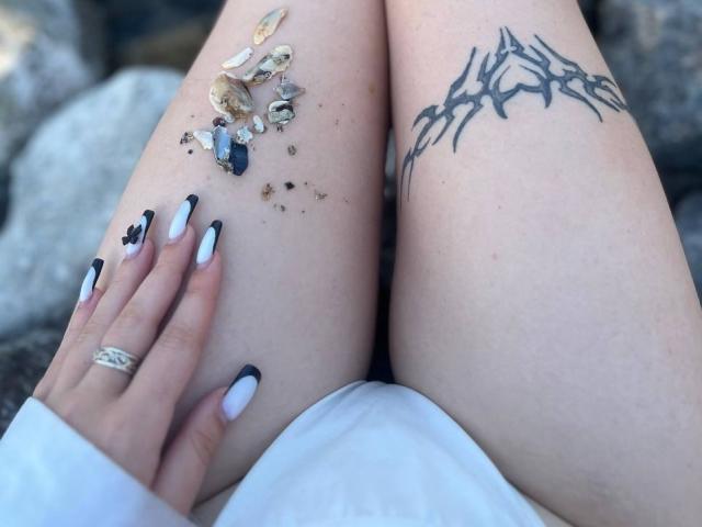 Las muslos pálidos de LaPulia, con tatuajes tribales negros, sus manos con esmalte de uñas negro y blanco, y un anillo, sosteniendo conchas marinas, sentada sobre rocas. El trasero de LaPulia es visible..