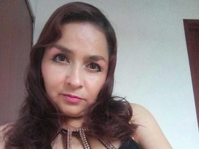 YaraStone tiene una piel marrón media, cabello ondulado de color marrón oscuro y lápiz labial rosa. Lleva un top negro con tirantes y un collar de cuentas, y está mirando a la cámara..