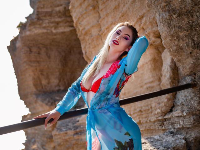 LindaBryns with blonde hair, red bikini top, and blue sheer robe, leaning against a metal railing, showing her boobs, standing against rocky cliffs, looking sexy.