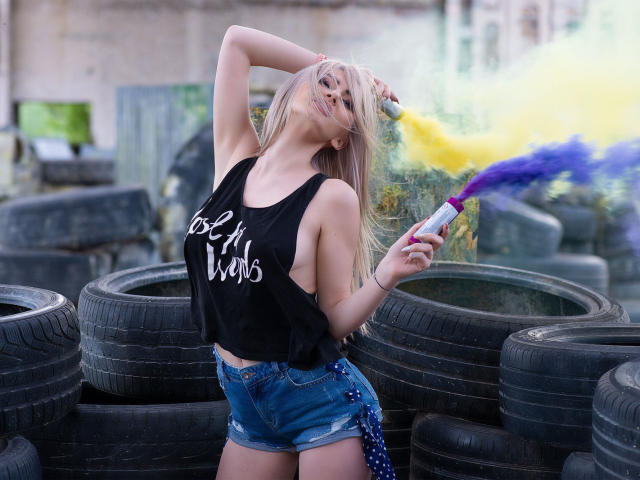 LindaBryns with blonde hair, black tank top showing her tits, blue shorts, spraying yellow and purple paint, standing among stacked tires, arm raised, looking at camera.