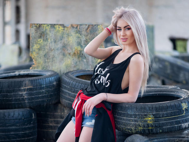 LindaBryns with blonde hair, black tank top, red shirt tied around waist, denim shorts, leaning on black tires, smiling, right hand in hair, left arm on tire.