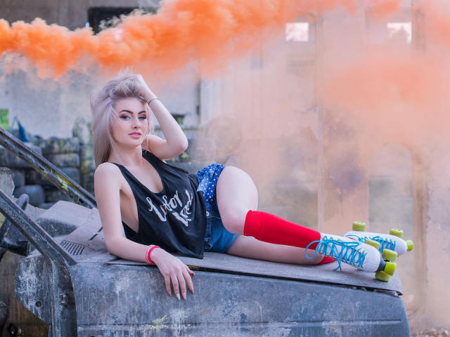 LindaBryns lying on a gray metal object, wearing a black tank top, blue shorts, red knee socks, and green roller skates, with orange smoke in the background.