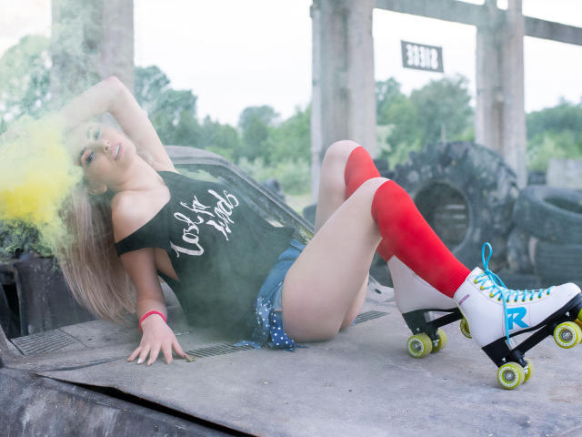 LindaBryns with blonde hair, black off-shoulder top, blue shorts, red knee socks, white roller skates, and yellow smoke, leaning back on a roller skate ramp.