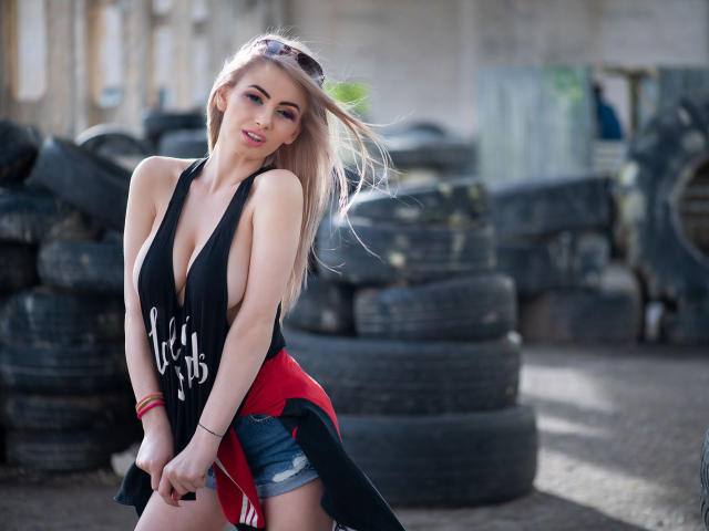 LindaBryns with big tits, wearing a low-cut black and red top, denim shorts, and sunglasses on head, standing in front of stacked tires. She's looking at the camera.