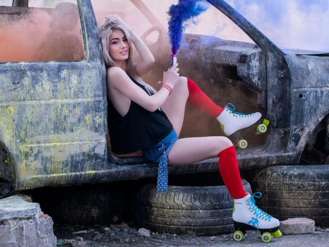 LindaBryns with blonde hair, black tank top, blue shorts, red knee socks, white roller skates, and blue smoke, sitting in a rusty, burnt-out car. Boobs visible.