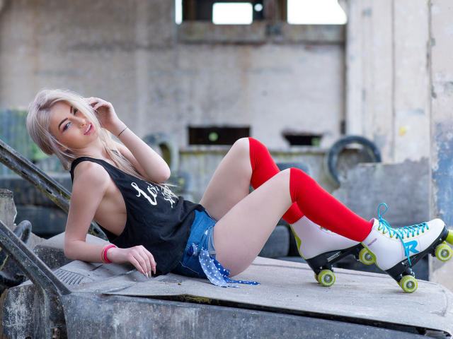 LindaBryns with platinum blonde hair, wearing a black tank top, red knee socks, blue denim shorts, and white roller skates with green wheels, sitting on concrete steps.