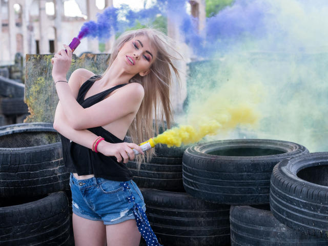 LindaBryns with blonde hair, black tank top, denim shorts, and red bracelet, holding purple and yellow smoke bombs, smiling, standing in front of stacked black tires.