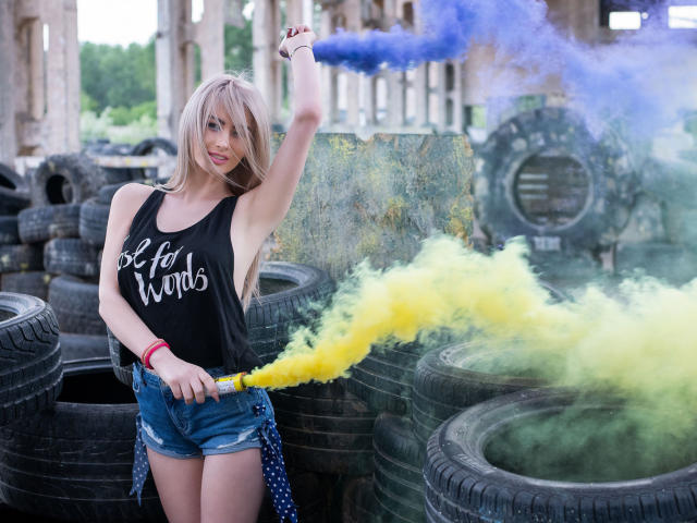 LindaBryns with blonde hair, black tank top, and denim shorts, holding yellow and blue smoke, standing among stacked tires. Her arm is raised, showing a red bracelet.