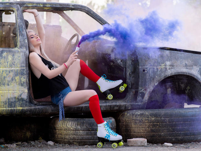 LindaBryns with blonde hair, red knee socks, white roller skates, and blue laces, sprays blue smoke from a bottle, sitting in a rusty, abandoned car.