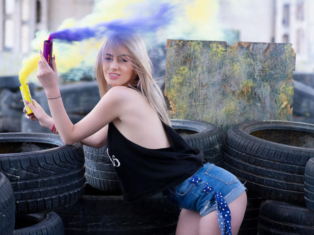 LindaBryns with blonde hair, smiling, wearing a black tank top and denim shorts, spraying yellow and blue paint, standing among black tires, holding a purple paint can.