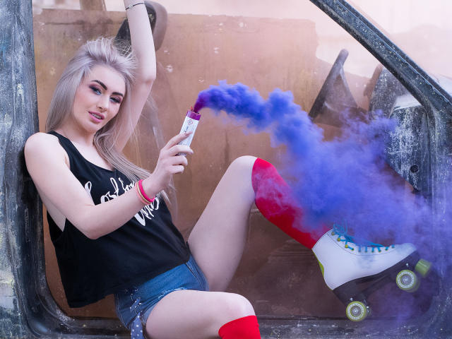 LindaBryns with blonde hair, black tank top, denim shorts, red knee socks, and white roller skates, blowing blue smoke from a vape pen, arm raised, sitting in a car.