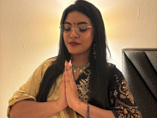 LakshmIrani, dark-haired, wearing glasses, and a black/gold patterned traditional outfit, with hands in prayer, has a calm expression, and is adorned with a necklace and earrings.