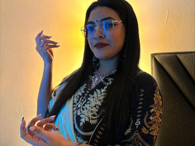 LakshmIrani, with long black hair, wearing a dark blue and gold embroidered top, and glasses, has her hand raised, showing dark nail polish, and a confident smile.