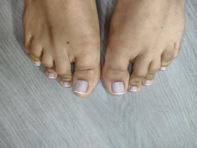 DanikaSerov's toes, showing light brown skin with a small mole on her left big toe, and white-painted toenails on a textured gray surface.