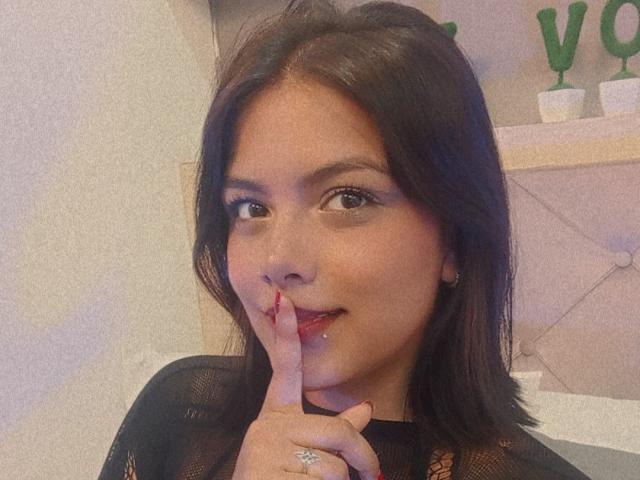 EmilyRobeerts with dark brown hair, wearing a black mesh top, has her finger to her red-painted lips, showing a subtle smile. She has a ring on her index finger.