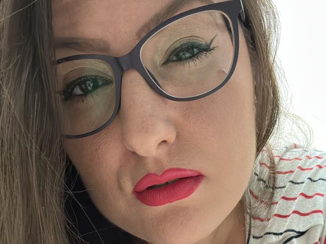 BadAngel's face with green eyeshadow, thick black eyelashes, red lipstick, and large black glasses. She has a small beauty mark near her right eye and a striped shirt.