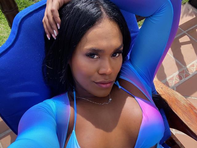 AshleyRob with dark skin, black hair, and blue-purple top, showing big tits, lying back in blue chair, arm raised, nail polish visible, outdoor, sunny, necklace, confident smile.