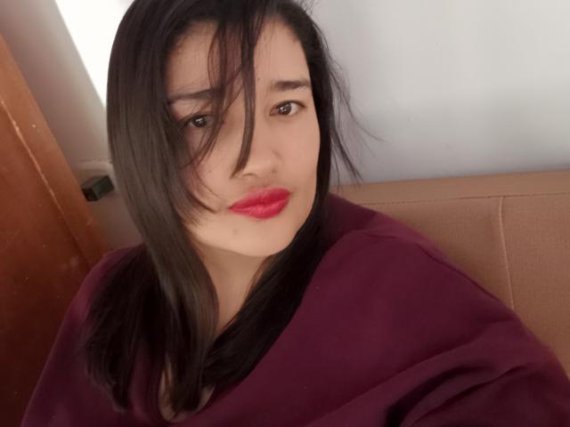 ZafiRouge with dark, straight hair, red lipstick, and a purple shirt, looking at the camera with a slight smile. Her tits are partially visible under the shirt.