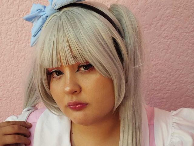 SpacyBabe69 ha i capelli cosplay grigio chiaro, con due trecce decorate da un fiocco blu, indossa bretelle rosa sopra una maglietta bianca e ha il rossetto rosa sulle sue guance leggermente paffute..