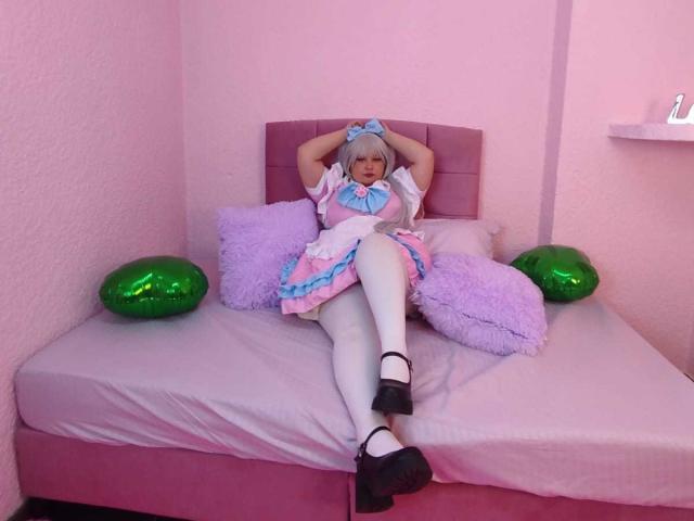 SpacyBabe69 with pink hair, blue bow, pink and white dress, white tights, black Mary Jane shoes, lying on pink bed, hands behind head, green and purple pillows.