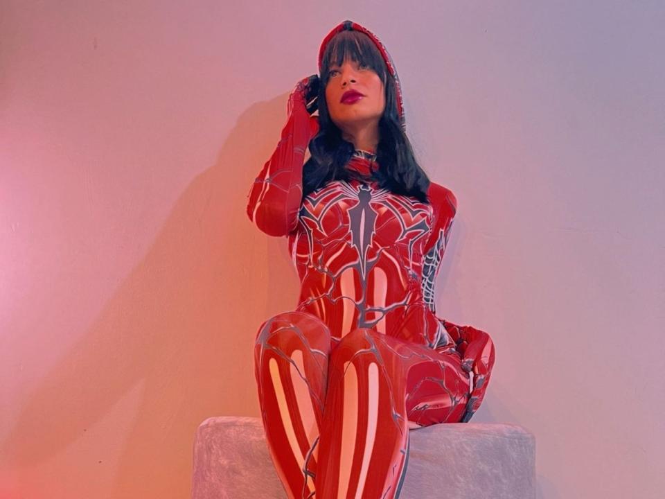 Varoneza in a red and white spider web-patterned latex outfit, sitting on a gray block, hand in her hair, with red lipstick and black bangs, looking seductive.