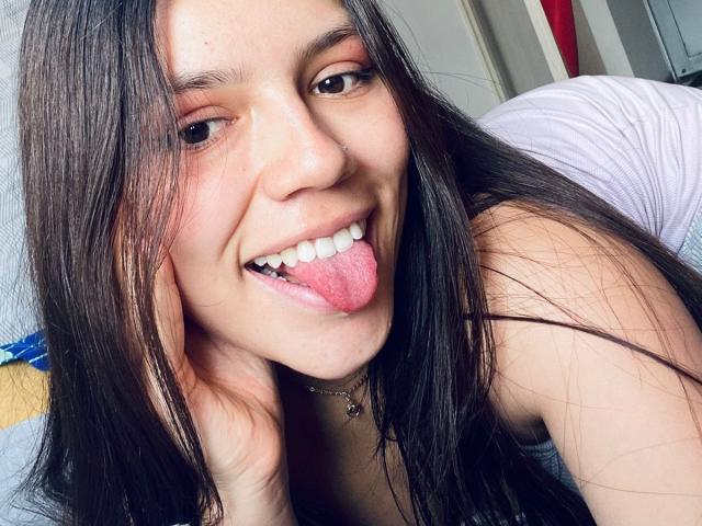 SophiaMarx with long brown hair, brown eyes, and fair skin, sticking out her pink tongue, smiling, and resting her head on her hand, wearing a white top.
