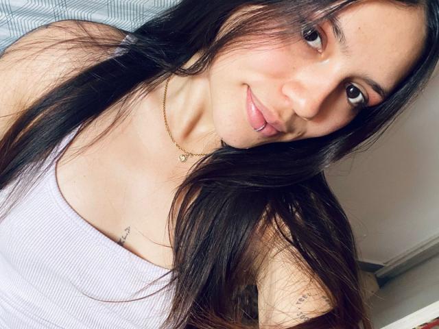 SophiaMarx, con cabello negro largo, sonriendo, pintándose los labios de rosa, vistiendo una camiseta blanca de tirantes con textura, un collar dorado y tatuajes sutiles en su brazo derecho..