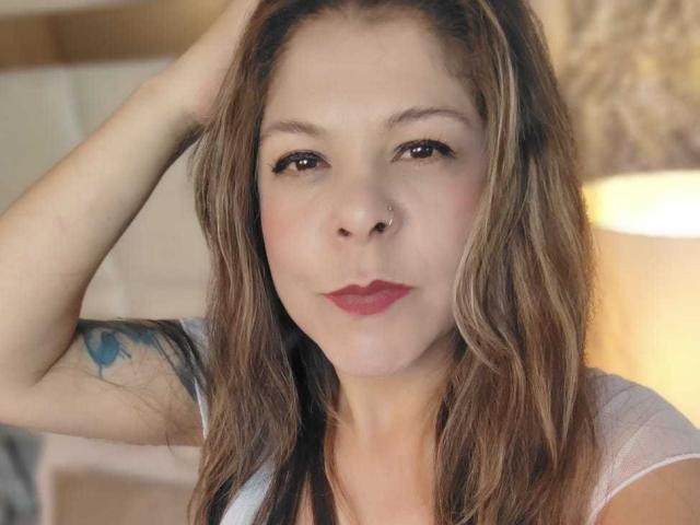 MartinaFiore hat braunes, welliges Haar, einen Nasenring, roten Lippenstift und ein Tattoo auf ihrem linken Arm. Sie schaut in die Kamera und hat eine Hand in ihrem Haar..