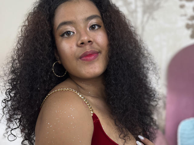 Adeleine Pierce, con cabello oscuro y rizado, vestida con una blusa de terciopelo rojo con tirantes de cadena dorada, aros grandes y lápiz labial rosa, mirando a la cámara con una ligera sonrisa..