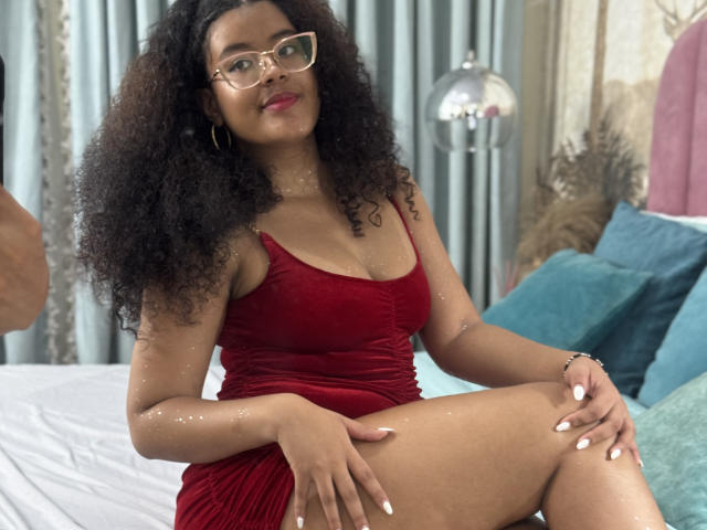 Adeleine Pierce, avec ses cheveux noirs bouclés, portant une robe en velours rouge, des lunettes et des boucles d'oreilles cercles, assise sur un lit, montrant ses seins, avec du vernis à ongles blanc et du rouge à lèvres rose..
