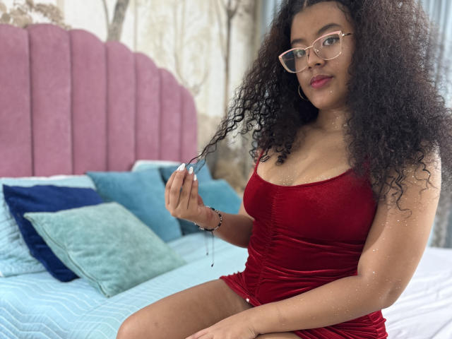 Adeleine Pierce, con i capelli neri ricci, indossa un abito di velluto rosso, si tiene i capelli, porta gli occhiali, è seduta sul letto con cuscini blu e verdi, una testiera rosa e una lenzuola blu..