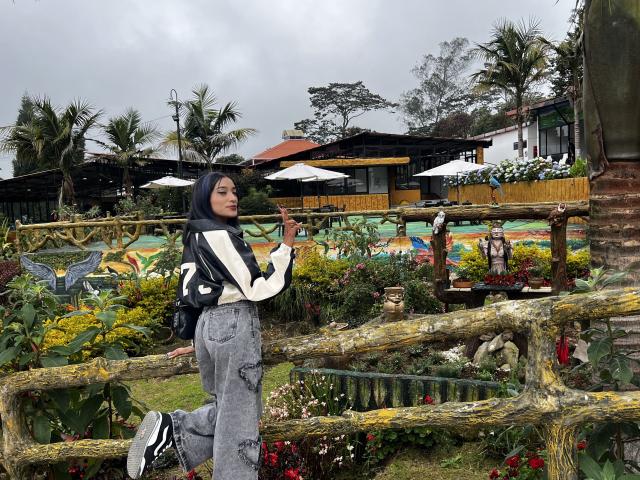 SalomeMille with black hijab, white long-sleeve shirt, grey cargo pants, black sneakers, standing by rustic wooden fence, holding cigarette, looking to the side, overcast sky.