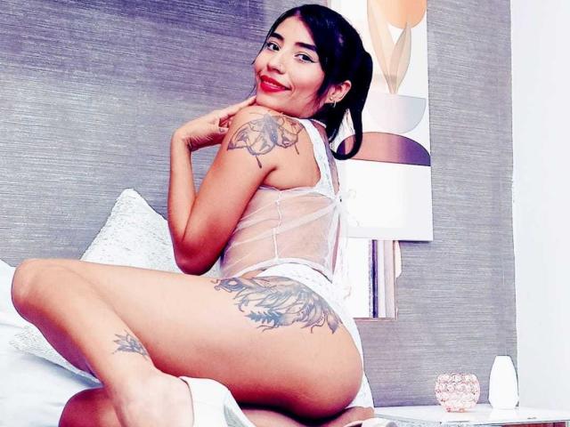 SexyRedLady with dark hair, red lipstick, white lace lingerie, sitting on bed, showing tattoo on thigh and arm, smiling, touching chin, revealing her ass and small tits.