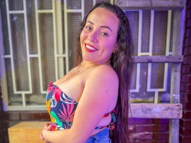 HoneyHotX with long dark hair, smiling, wearing a colorful floral strapless top, showing her tits, and blue jeans, standing in a slightly purple-lit room.