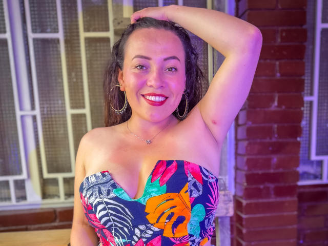 HoneyHotX con grandes senos, vestido sin tirantes de color floral, lápiz labial rojo, sonriendo, brazo levantado, aros grandes, collar, cabello castaño, luz rosa-morada, de pie frente a una pared de ladrillo..