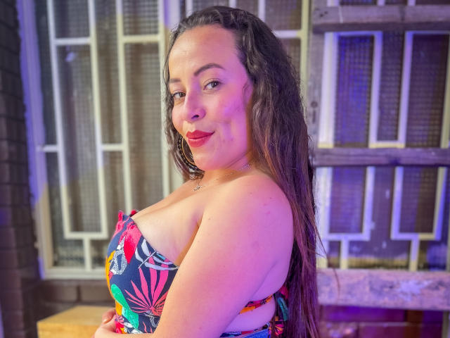 HoneyHotX con cabello largo y oscuro, labios rojos y un vestido tropical colorido sin tirantes, mostrando sus grandes senos y sonriendo a la cámara. El fondo está desenfocado..