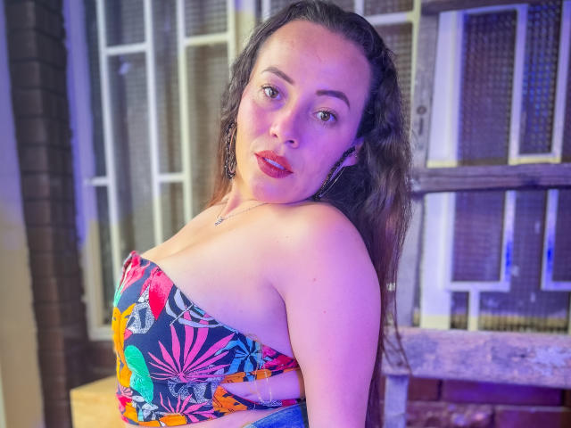 HoneyHotX, con i capelli scuri, il rossetto rosso e il top senza spalline con motivi floreali colorati, che mette in mostra il seno, guarda la fotocamera con un leggero sorriso. Lo sfondo è sfocato..