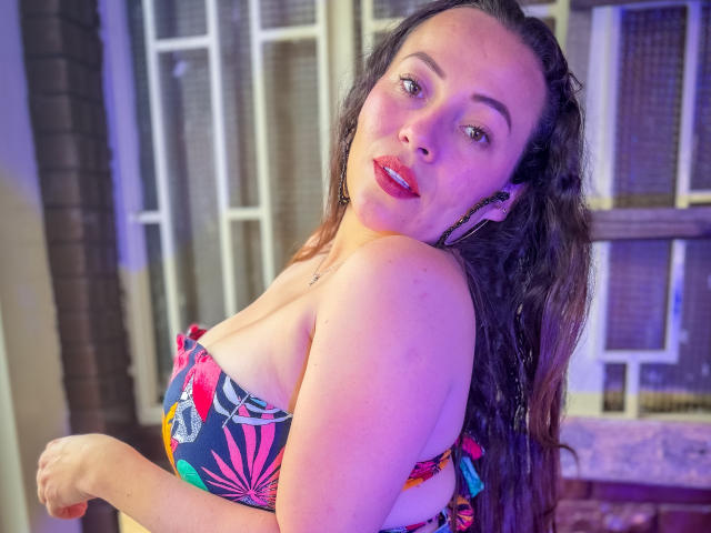 HoneyHotX mit feuchtem, braunem Haar, rotem Lippenstift, trägt einen bunten, blumigen Bikini, zeigt ihre Brüste, hört mit Kopfhörern Musik, schaut mit leicht geöffnetem Mund zurück, in einem violett beleuchteten Raum..