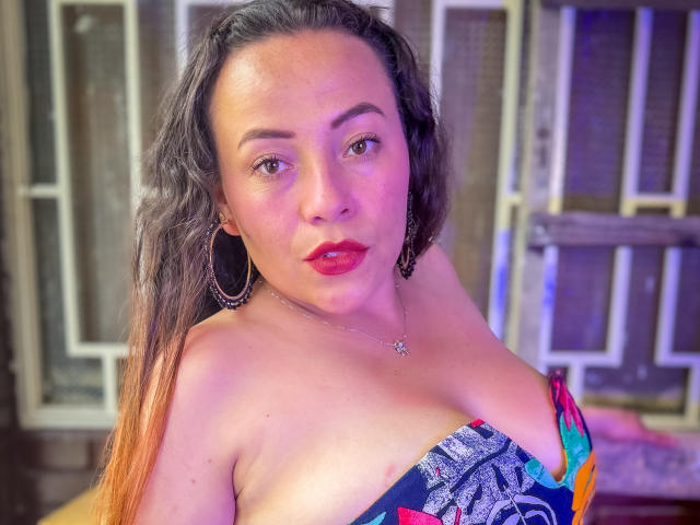 HoneyHotX, con cabello largo y oscuro, lápiz labial rojo, aros grandes y una blusa colorida con estampado que muestra un busto generoso, está de pie en un interior con iluminación púrpura..