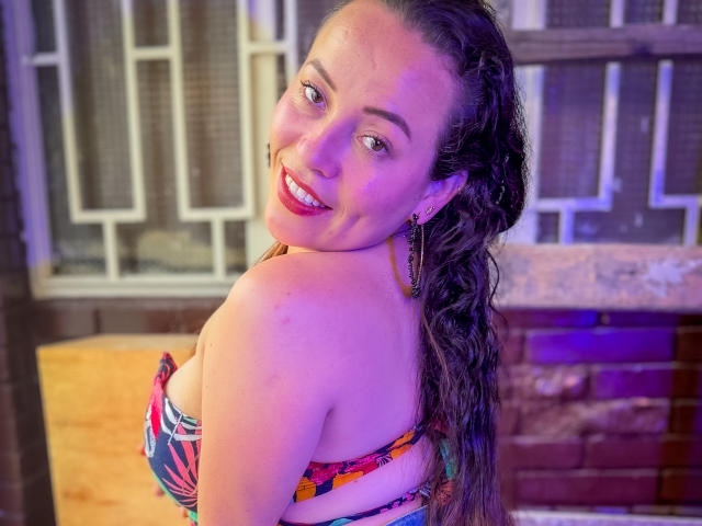 HoneyHotX with wet brown hair, smiling, showing her tits in a colorful strapless top, purple-blue lights, hoop earrings, and red lipstick, looking over her shoulder.