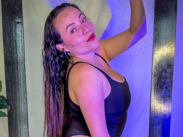 HoneyHotX with wet long brown hair, red lipstick, black tank top, showing her ass, arm raised, looking at camera, blue and purple light, earrings, smiling.