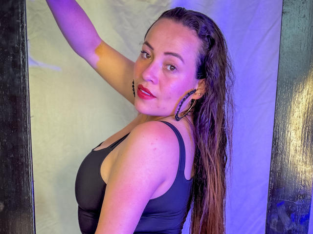 HoneyHotX with wet brown hair, black tank top, red lipstick, hoop earrings, looking over shoulder, smiling, showing her ass, in a dimly lit, purple-hued setting.