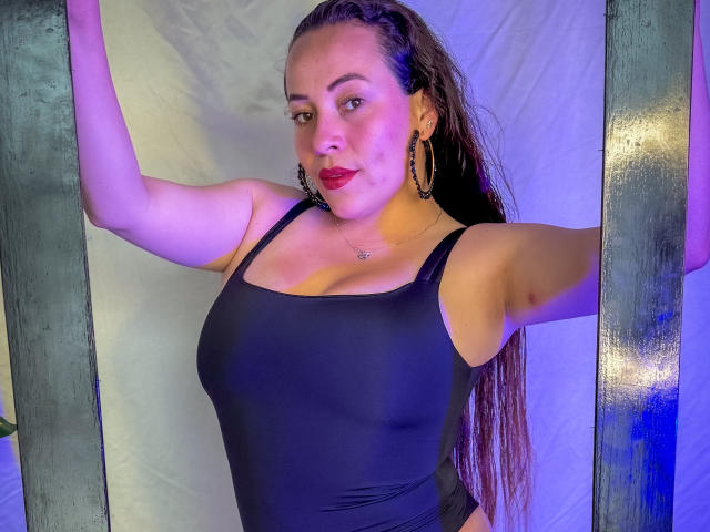 HoneyHotX with wet brown hair, wearing a black one-piece, big tits, and gold hoop earrings, arms raised, red lipstick, and a slight pout, standing between green wooden beams.