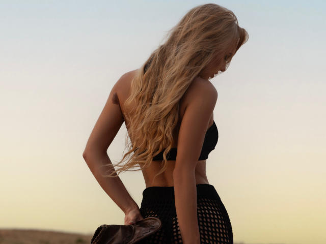 FloraDavis with long blonde hair, black bra, and black mesh skirt, holding brown bag, standing sideways, showing her ass and back, under a clear sky.