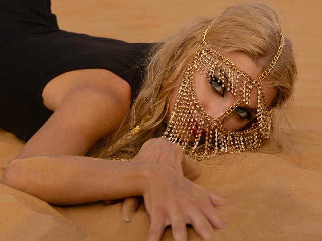 FloraDavis is lying in the sand with her face partially covered by a gold chain mask, showing her tits and a serious expression, wearing a black top.